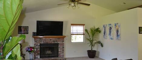 A living room with a brick fireplace, a flat-screen TV, a ceiling fan, and a large potted plant.