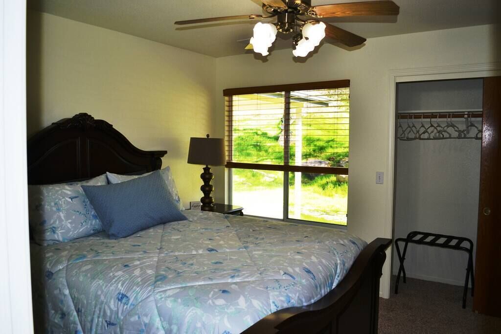 Une chambre à coucher avec un lit, un ventilateur de plafond, une fenêtre avec des stores et une porte dotée d’un porte-manteau.