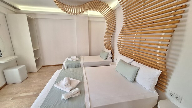 A modern hotel room with two beds, a wooden slat headboard, a small seating area, and a built-in TV. - Ergin Butik Otel (Bozcaada)
