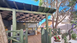 A covered outdoor seating area with wooden benches and a grill, surrounded by trees and a white wall. - Ergin Butik Otel (Bozcaada)