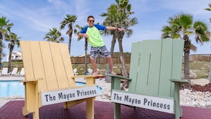 A person standing on two Adirondack chairs labeled "The Mayan Princess" with a pool and palm trees in the background. - Escape to Mayan Princess 213 Beachfront Getaway! (Port Aransas)