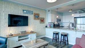 A modern kitchen with white cabinets, a stainless steel refrigerator, and a flat-screen TV mounted on the wall. - Escape to Mayan Princess 213 Beachfront Getaway! (Port Aransas)