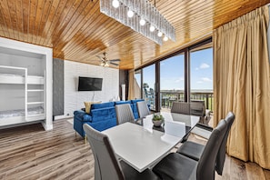 A modern living room with a wooden ceiling, a blue sofa, a dining table with chairs, and a large window with curtains.
