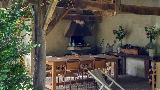 Un área de comedor al aire libre de estilo rústico, con una mesa y sillas de madera, rodeada de follaje y una chimenea.