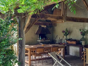 Un espace de restauration extérieur rustique, avec une table et des chaises en bois, entouré de verdure et agrémenté d’une cheminée.