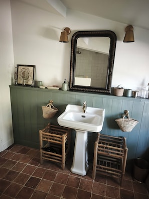 Ein Badezimmer mit einem weißen Waschbecken, einem großen Spiegel und einer grünen Kommode. Es befinden sich Weidenkörbe und ein Holzschemel dort.