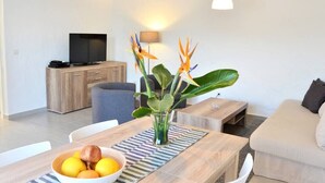 A modern living room with a wooden dining table, a bowl of fruit, a vase with flowers, a sofa, and a TV in the background. - Casa La Moringa - Holiday house close to the beach (Los Caideros)