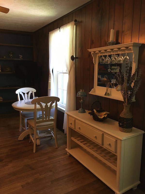 A wooden-paneled room with a dining table and chairs, a cabinet with glassware, and a window with curtains.