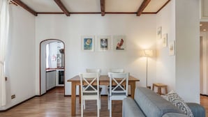 Una sala de estar con una mesa de comedor de madera, dos sillas blancas, un sofá gris y una cocina al fondo.