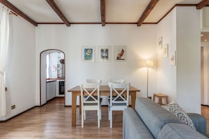 Uma sala de estar com uma mesa de jantar de madeira, duas cadeiras brancas, um sofá cinza e uma cozinha ao fundo.