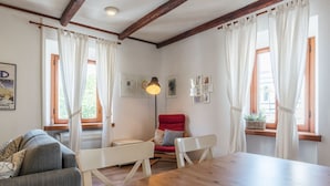 Una habitación con una mesa de madera, dos sillas blancas, una silla roja, un sofá y una ventana con cortinas.