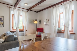 Quarto com mesa de madeira, duas cadeiras brancas, uma cadeira vermelha, um sofá e uma janela com cortinas.