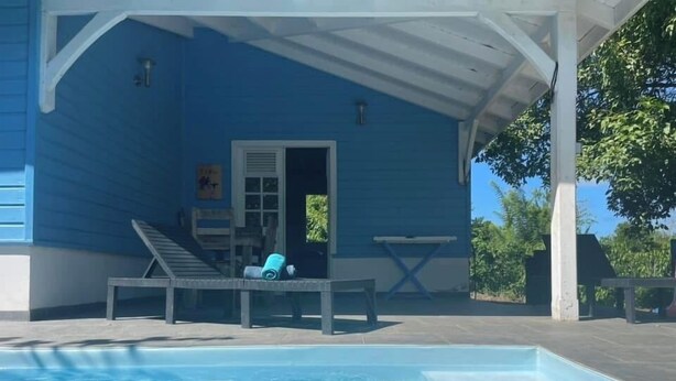 Een overdekt zwembadgedeelte met een blauw huis, ligstoelen en een kleine tafel.