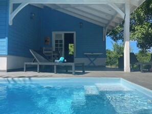 Zona de piscina exterior techada con una casa azul, sillas de descanso y una mesita.