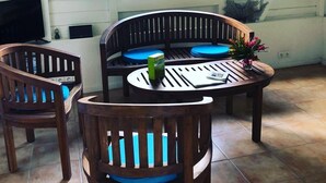 Een kamer met een houten tafel en stoelen, een raam met luiken, en uitzicht op groen buiten.