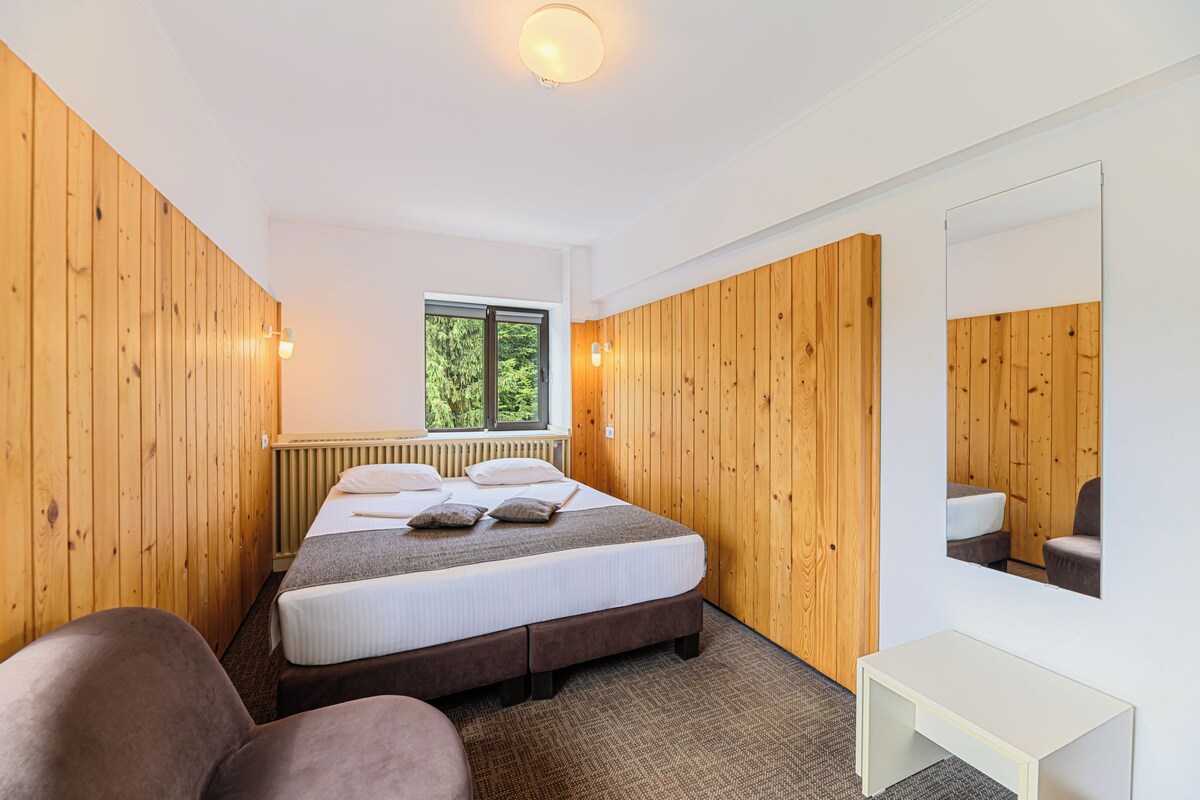 A bedroom with a bed, a window, a radiator, a mirror, and a small table.