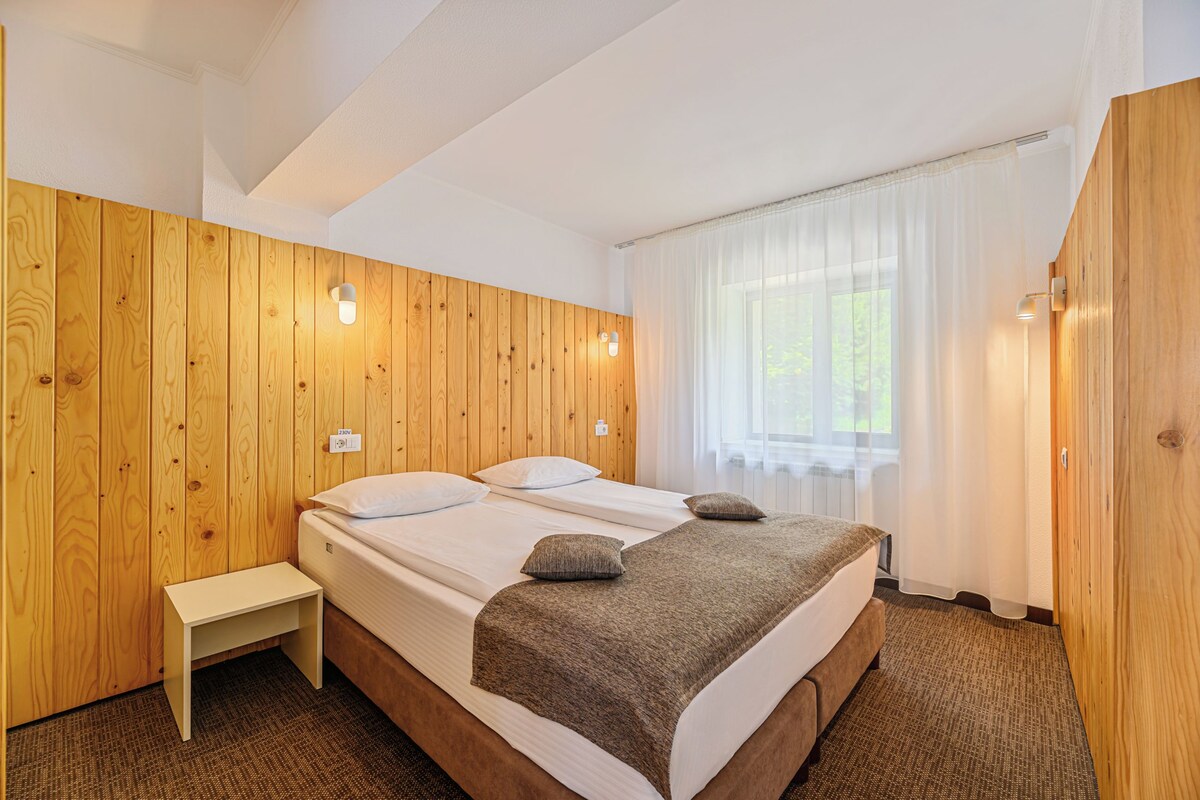 A hotel room with a wooden wall, a bed with white linens, a bedside table, and a window with curtains.