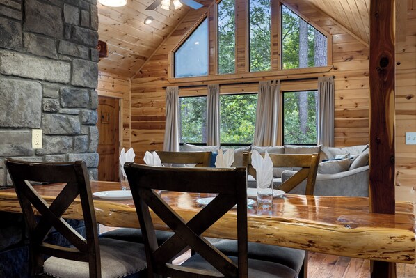A wooden cabin with a dining area featuring a wooden table and chairs, a stone wall, and large windows offering a view of the forest.