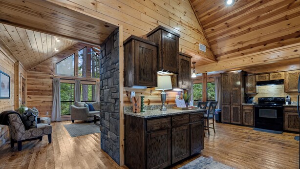 Ein Blockhaus-Interieur mit einer Wand aus Stein, Holzboden, einer Küche mit Herd und einer Sitzecke mit Sofa und Sesseln.