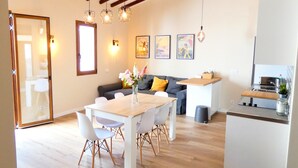 A modern kitchen with wooden beams, a dining area with a white table and chairs, and a living area with a sofa and cushions.