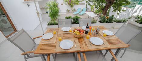 Un espace de restauration en plein air avec une table en bois dressée pour deux personnes, entourée de chaises, et une vue sur un jardin avec un hamac et un arbre.