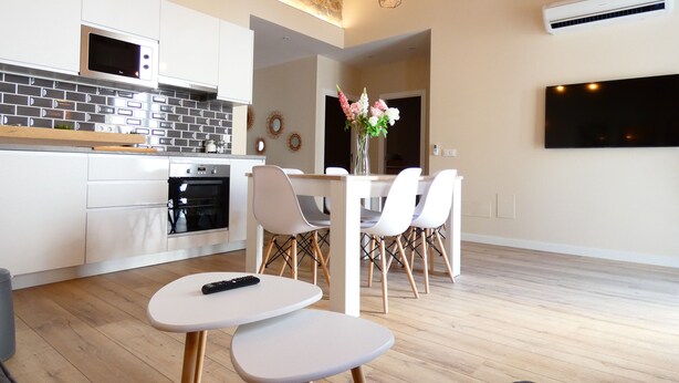 A modern kitchen with white cabinets, a microwave, and an oven. There is a dining area with a table and chairs, and a TV mounted on the wall.