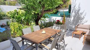 An outdoor patio with a wooden table and chairs, surrounded by lush greenery and a large tree.