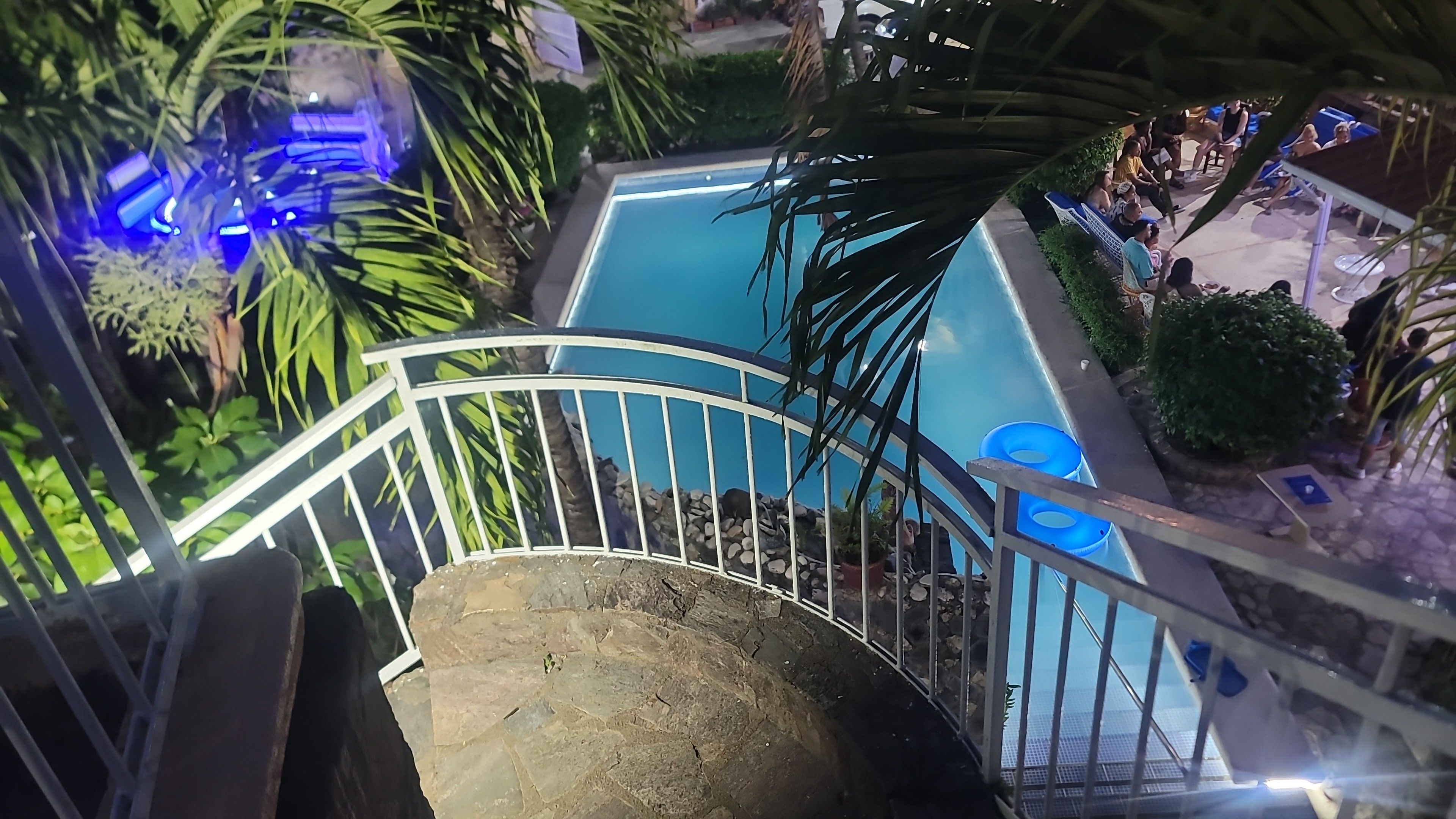 Zona de piscina con una escalera que baja hacia ella, rodeada de follaje y una barandilla.