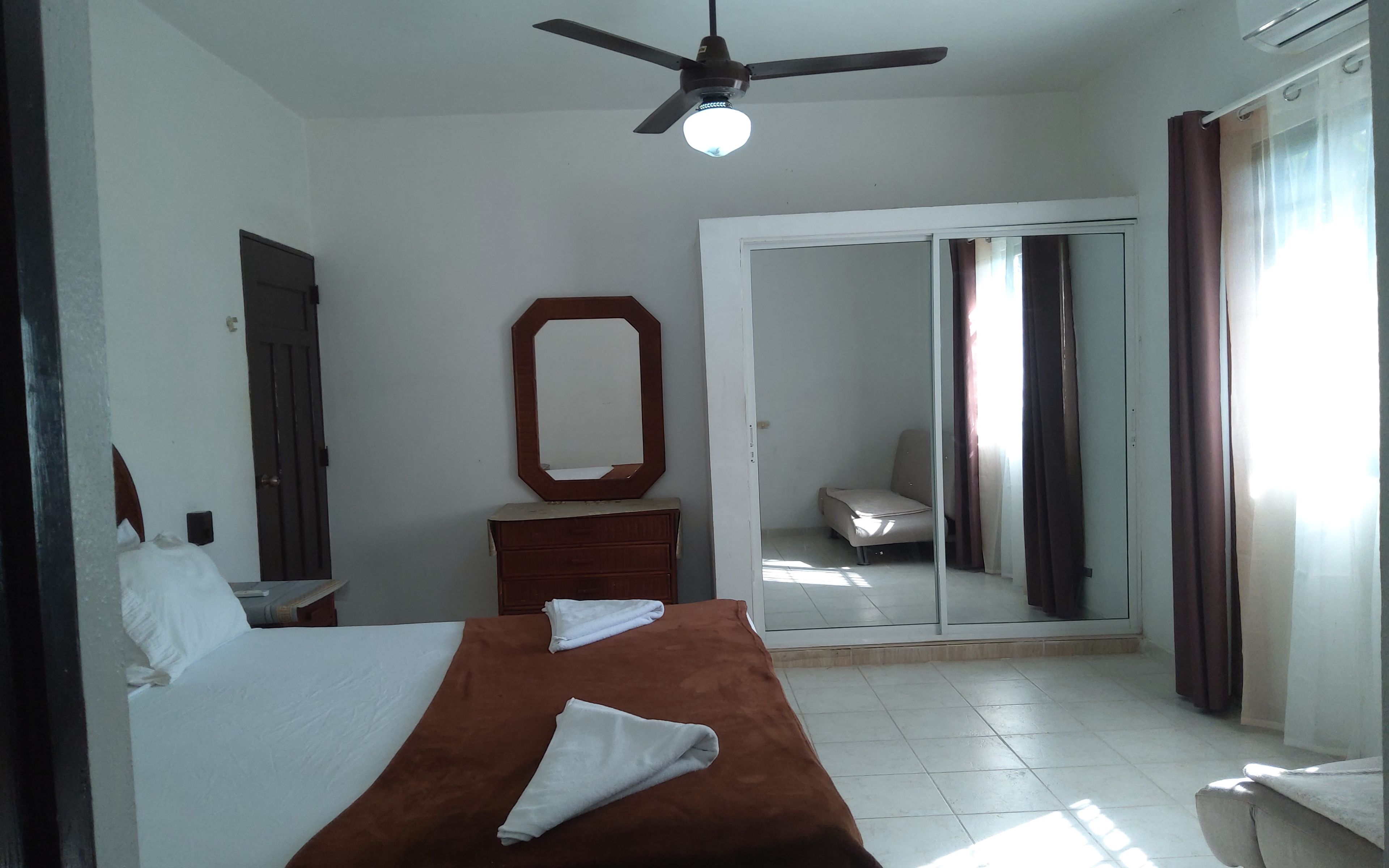 Habitación de hotel con cama, cómoda, espejo, ventilador de techo y puerta corrediza de vidrio que da a un balcón.