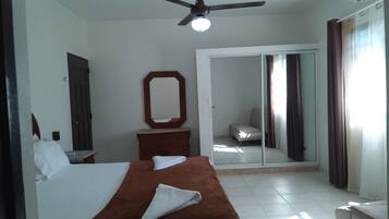 Habitación de hotel con cama, cómoda, espejo, ventilador de techo y puerta corrediza de vidrio que da a un balcón.