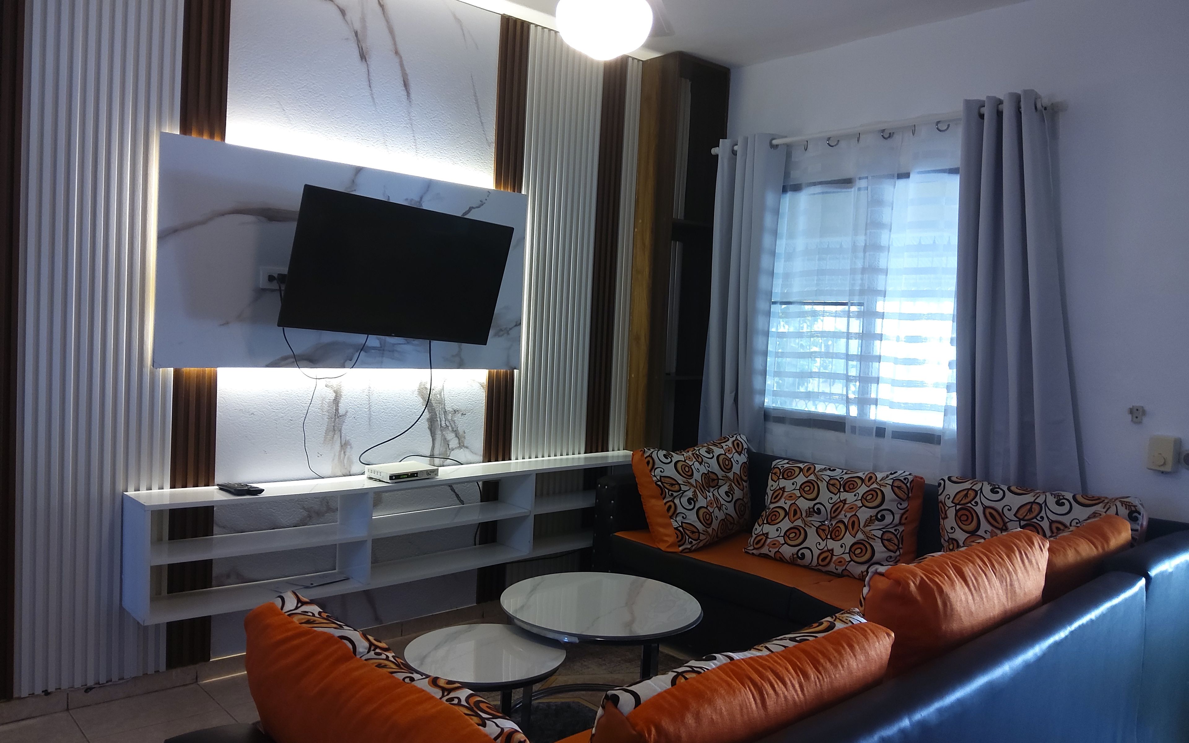 Una sala moderna con un televisor de pantalla plana montado en la pared, un sofá con cojines estampados, una mesita redonda y una ventana con cortinas.