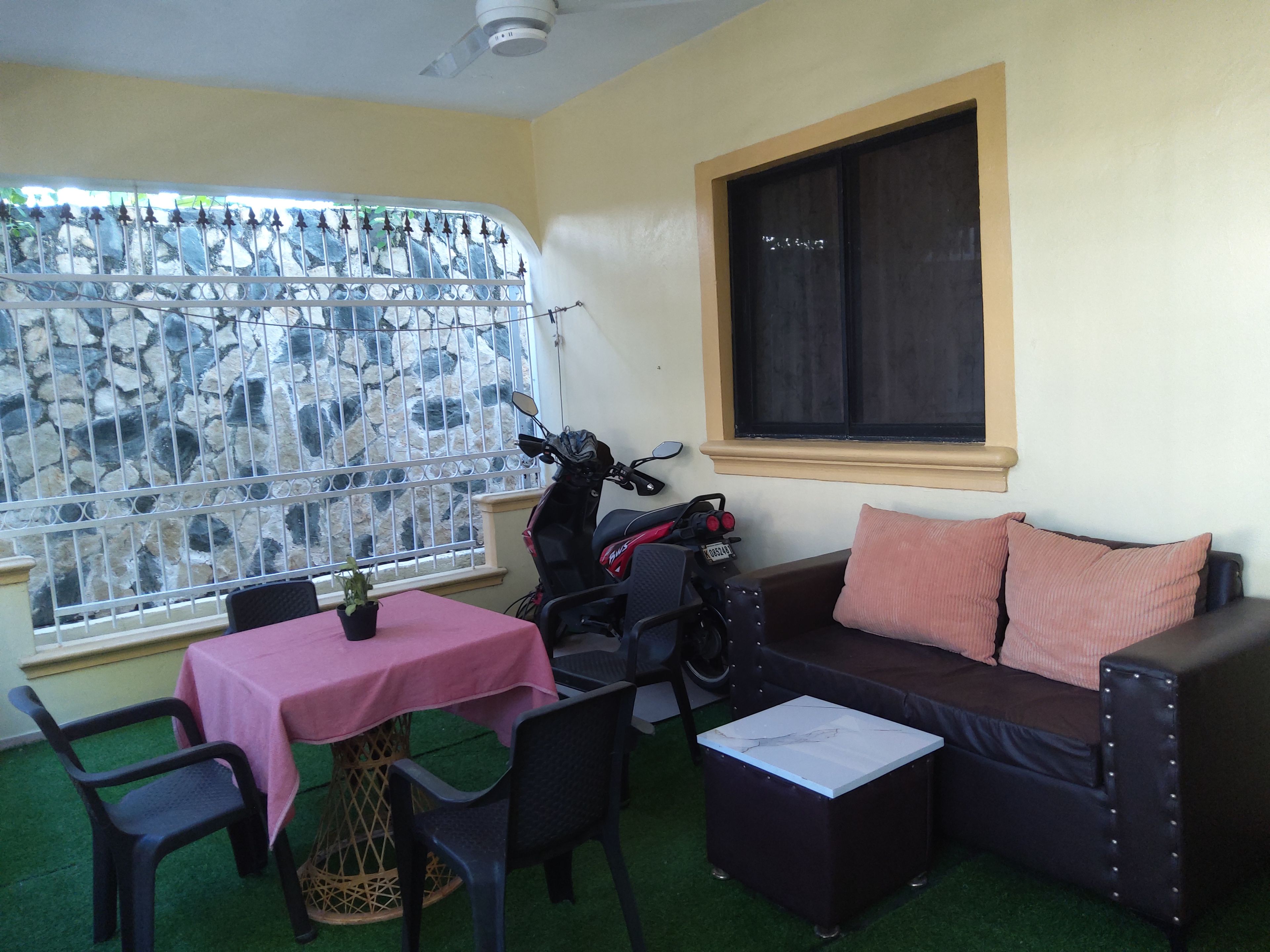 Una habitación con sofá, una mesa con mantel rosa, una motocicleta y una ventana con vista a un muro de piedra.