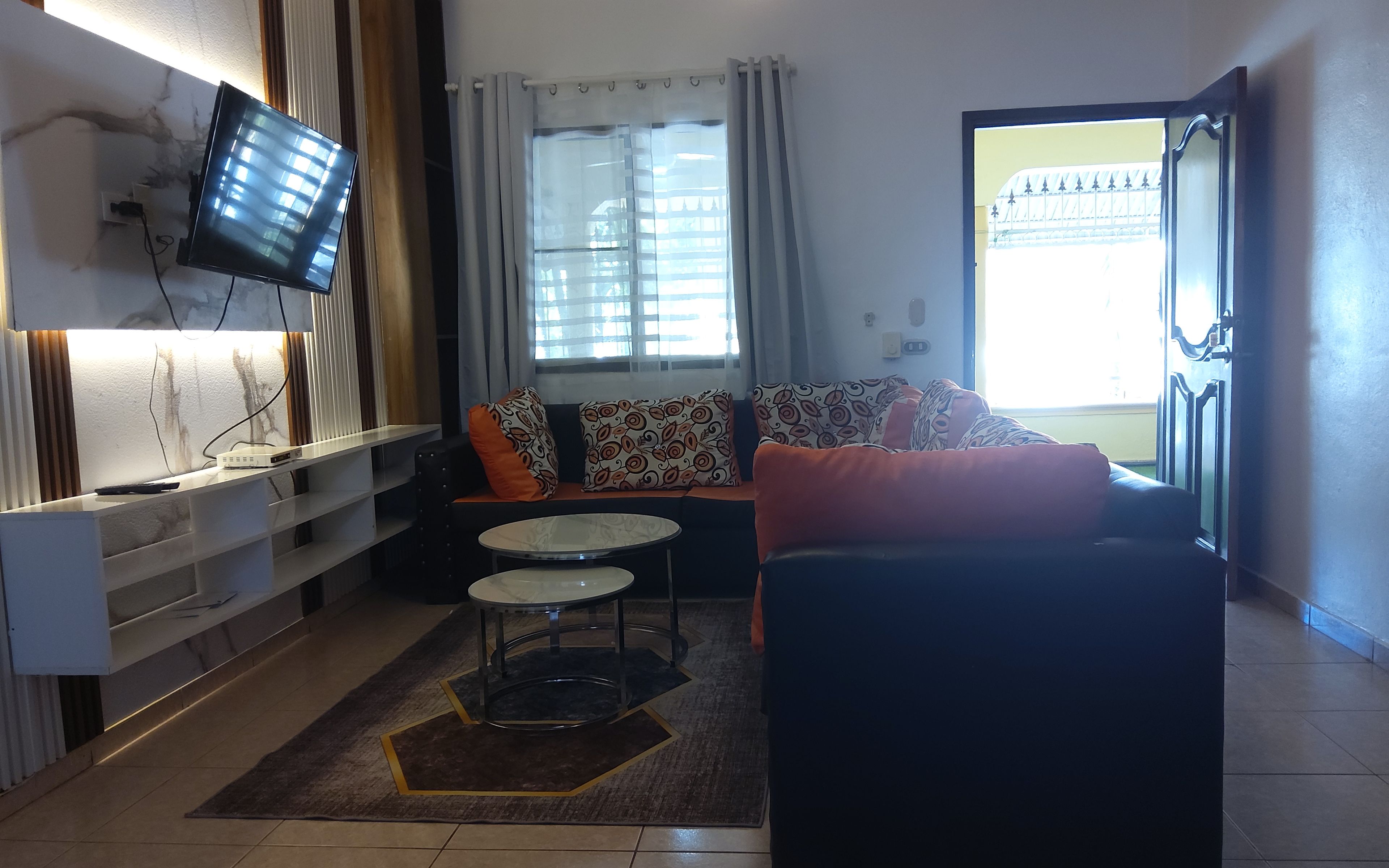 Un salon comprenant un canapé, une table basse, un téléviseur fixé au mur et une fenêtre avec des rideaux.