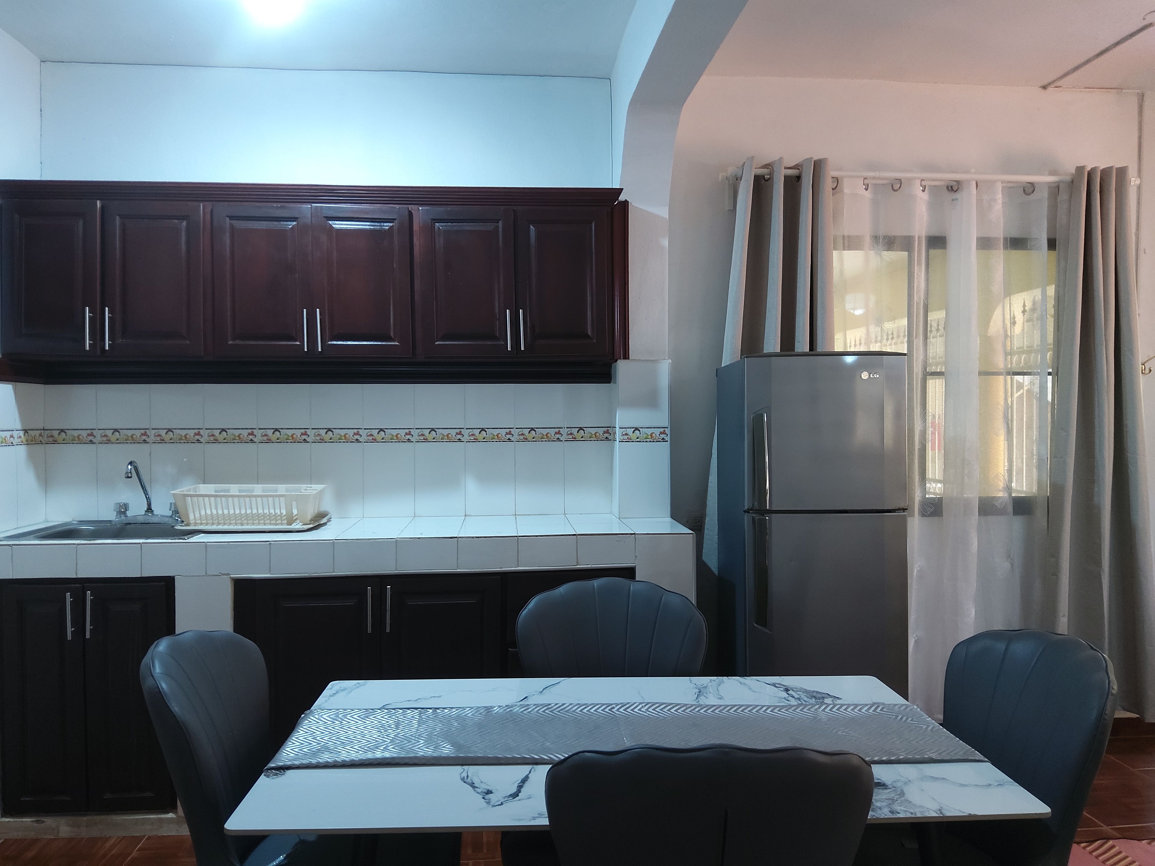 Una cocina con armarios de madera oscura, un refrigerador de acero inoxidable, una mesa de comedor con sillas y una ventana con cortinas translúcidas.