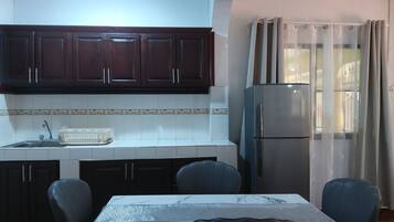 Una cocina con armarios de madera oscura, un refrigerador de acero inoxidable, una mesa de comedor con sillas y una ventana con cortinas translúcidas.
