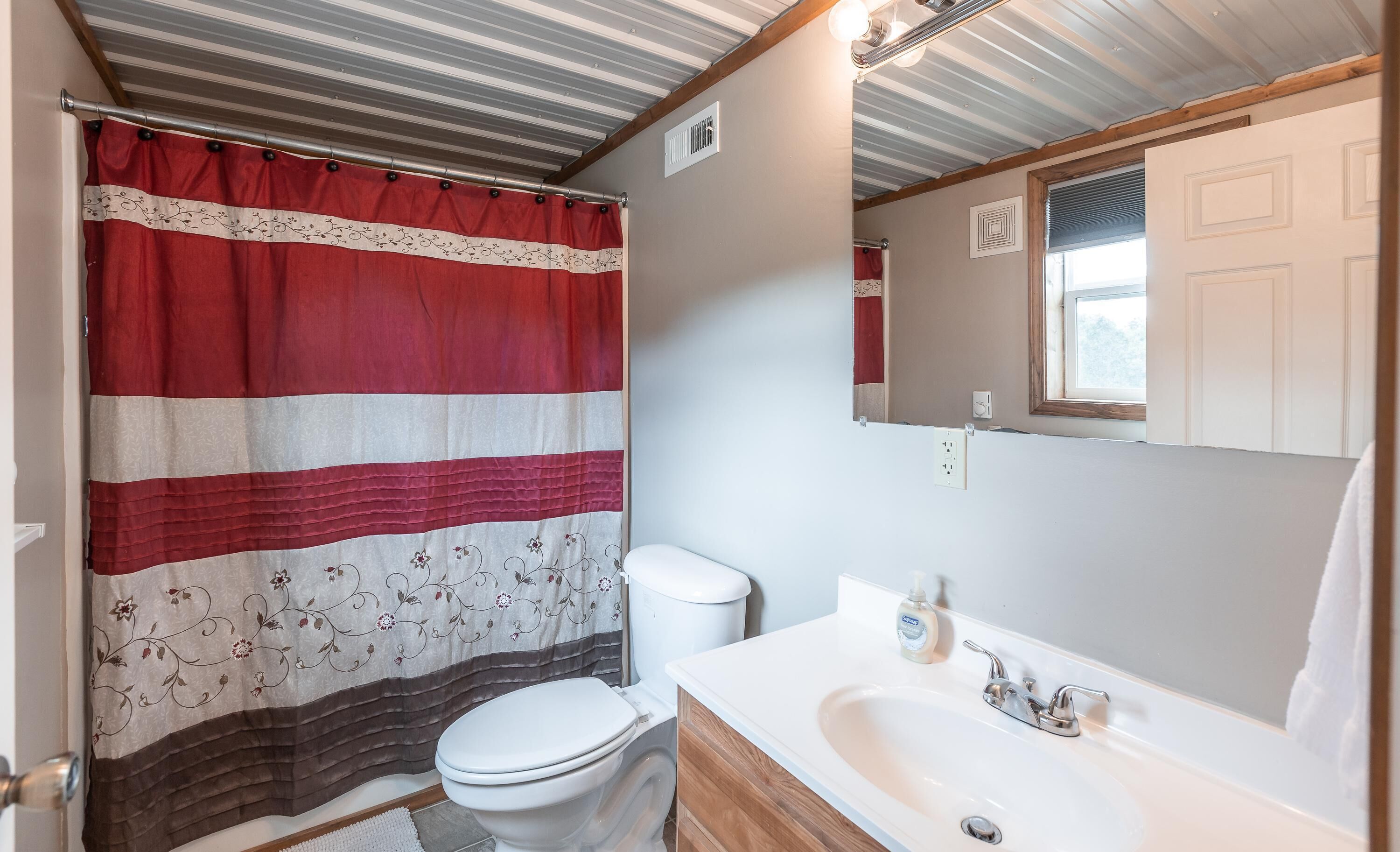 Une salle de bain avec un rideau de douche à rayures, des toilettes, un lavabo avec un miroir et une fenêtre avec des stores.