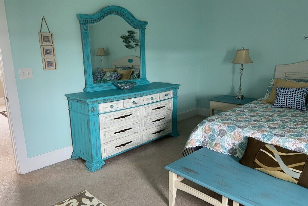 A bedroom with a turquoise dresser, a bed with patterned bedding, and a bedside table with a lamp.
