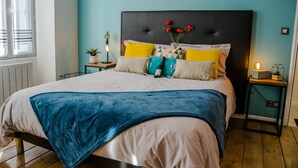 A neatly made bed with a blue blanket, a variety of pillows, and a vase with red flowers on a headboard. - BnB Epernay   Studio 86   Your Cozy Nest for Two in Épernay (Épernay)