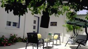 An outdoor seating area with metal chairs and a table, surrounded by white walls and a tree providing shade.