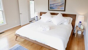 Een net opgemaakt bed met witte lakens, een houten hoofdbord en een schilderij erboven. Twee nachtkastjes met lampen en een klein vloerkleed op de vloer.