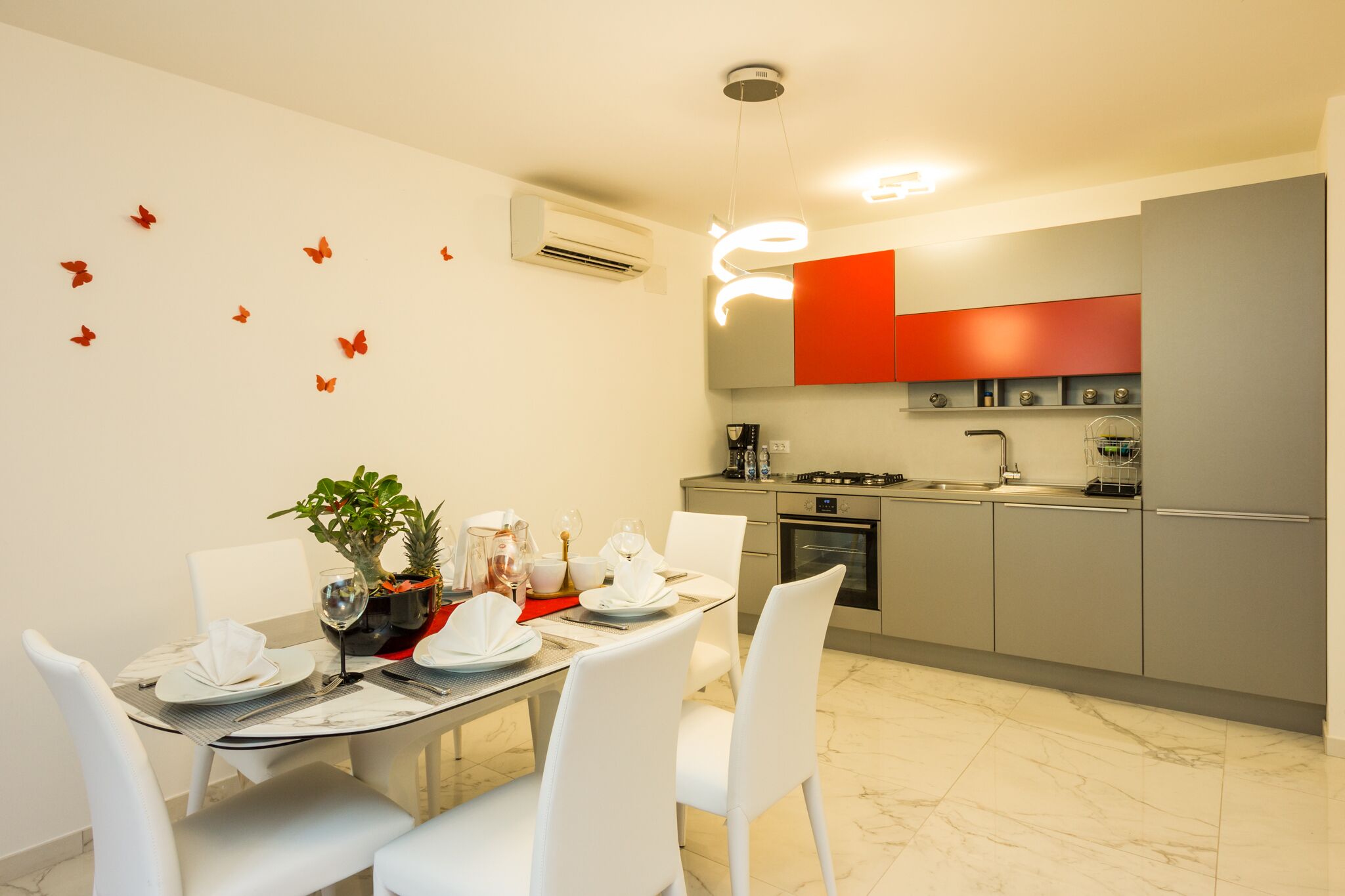 Una cocina moderna con armarios grises, una pared de acento roja y una zona de comedor con sillas blancas y una mesa de vidrio.