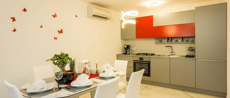 Una cocina moderna con armarios grises, una pared de acento roja y una zona de comedor con sillas blancas y una mesa de vidrio.