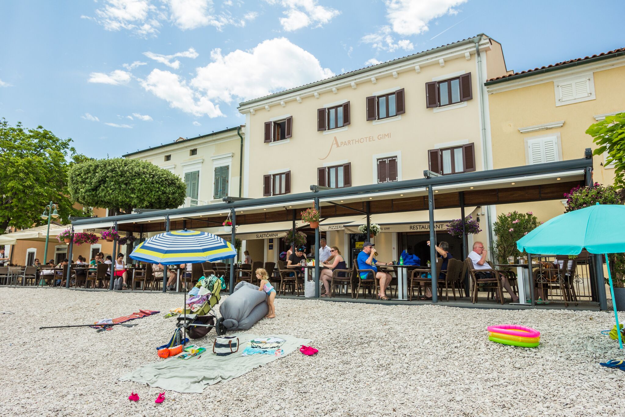 A beachside restaurant with outdoor seating, a covered area, and a building labeled "APARTHOTEL GIMI" in the background.