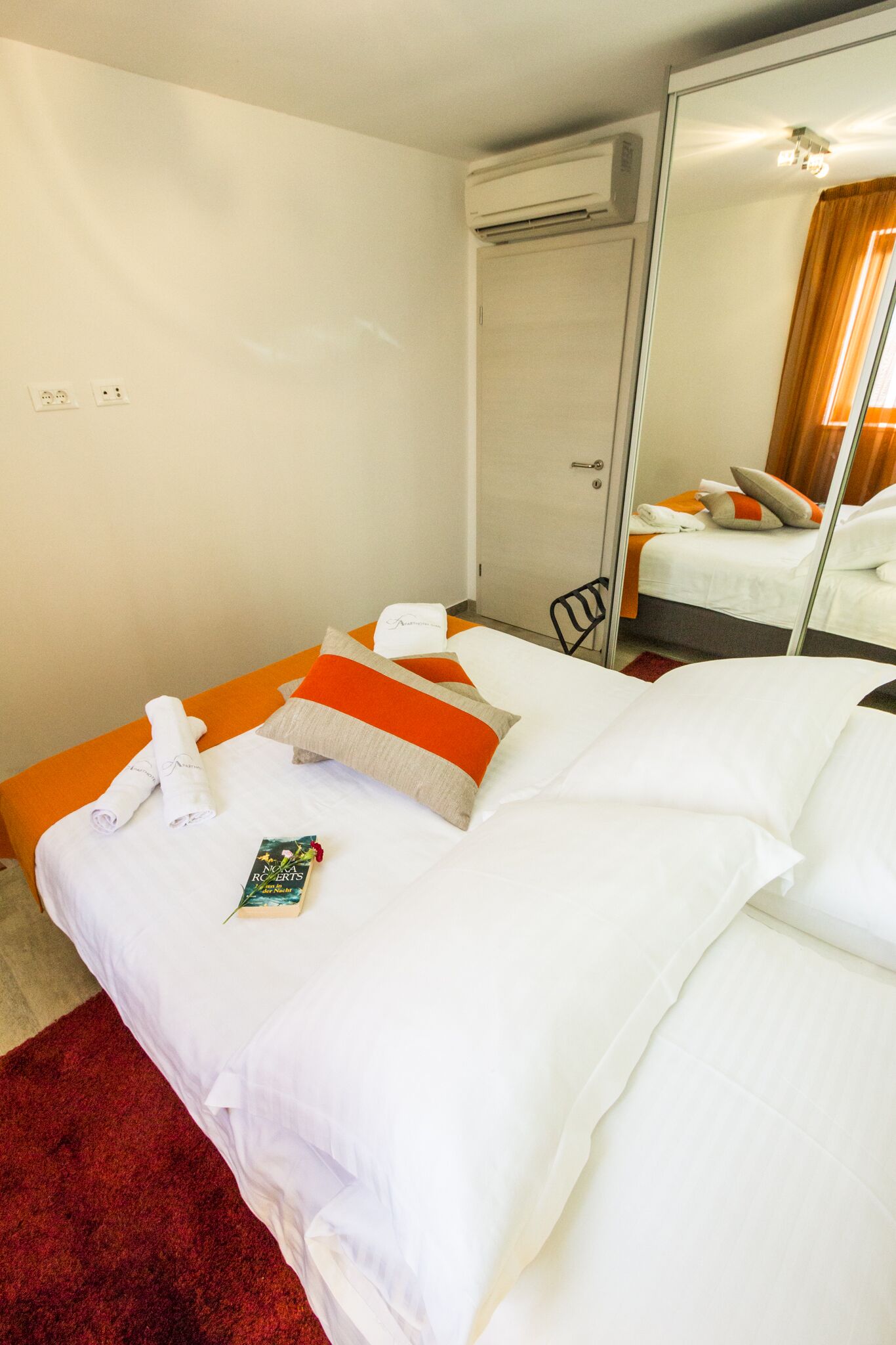 A hotel room with a bed, a mirror, an air conditioning unit, and a red carpet.
