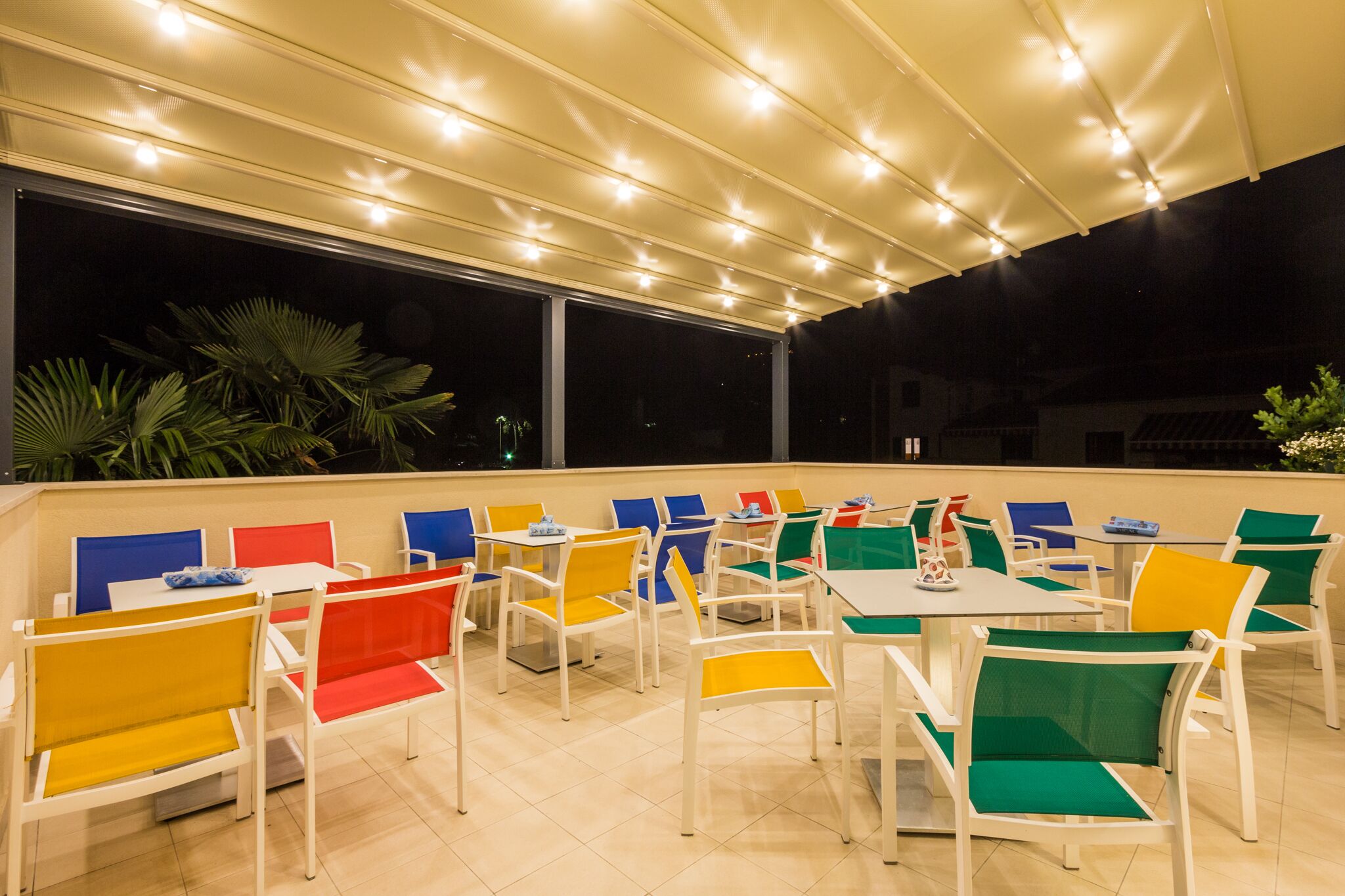 An outdoor dining area with white tables and chairs, some with colorful cushions.