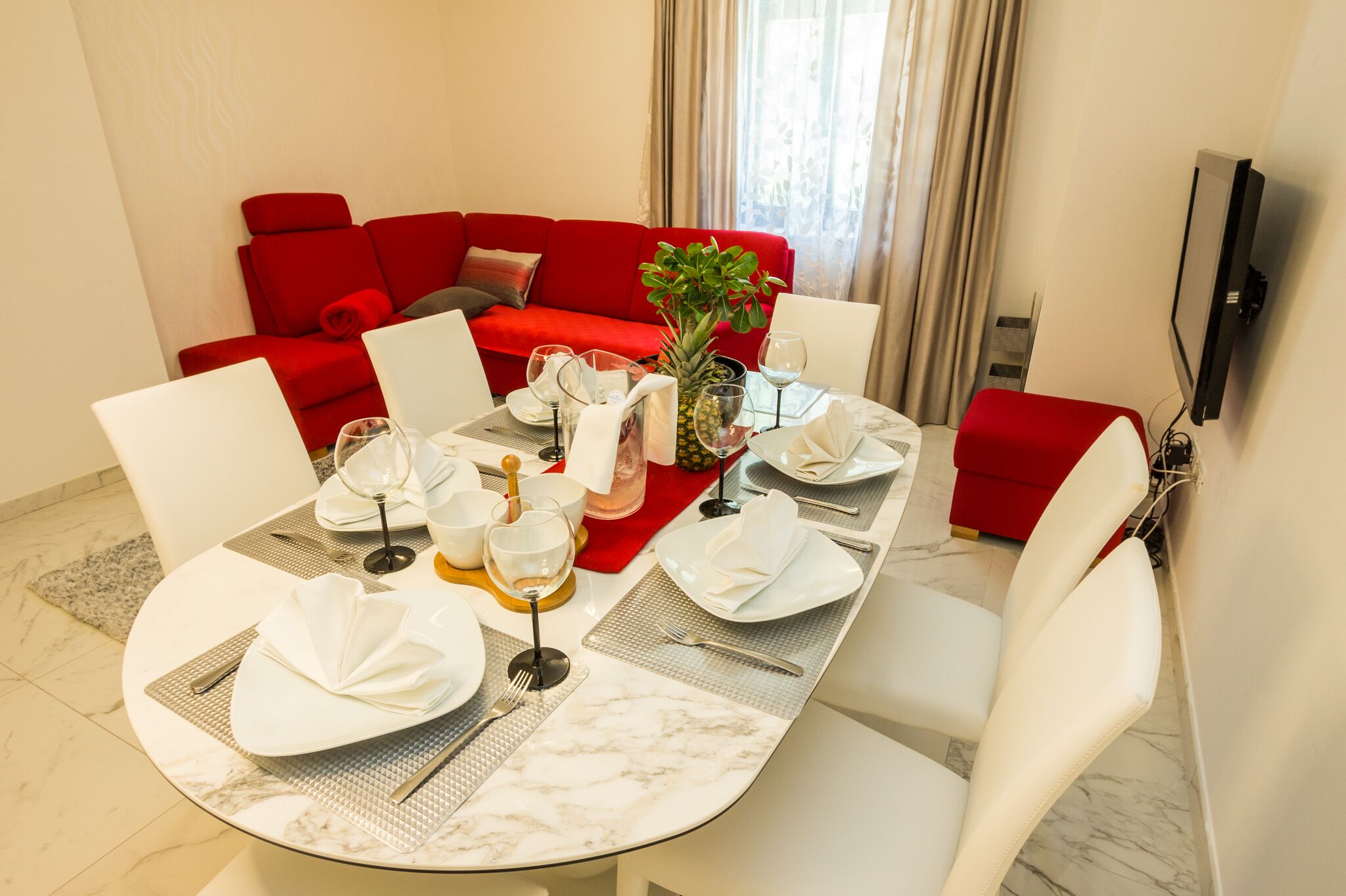 Un coin repas avec une table dressée pour quatre personnes, un canapé rouge en arrière-plan et un téléviseur fixé au mur.