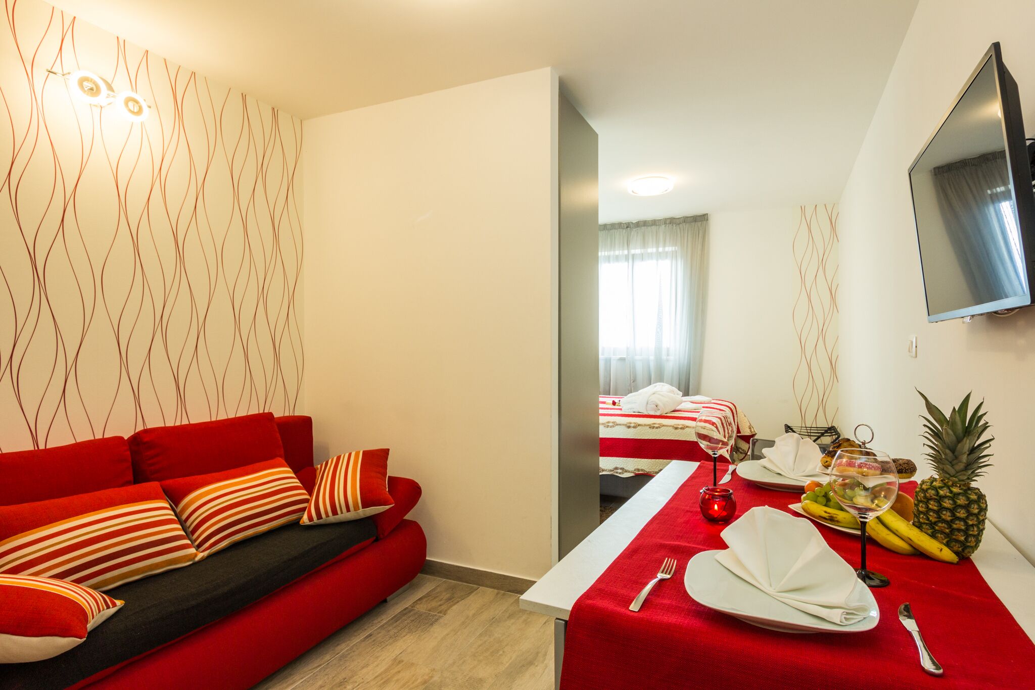 A modern hotel room with a red sofa, a dining table set with a red tablecloth, and a bed with white linens.