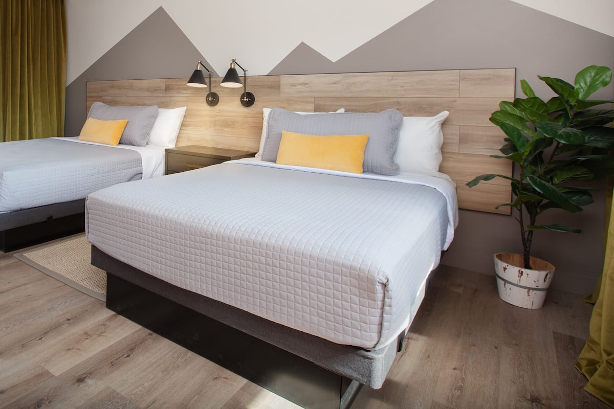 Une chambre d’hôtel avec deux lits, une tête de lit en bois, une plante en pot et une lampe fixée au mur.