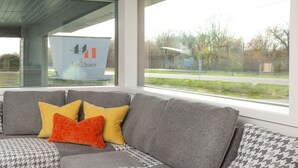 A modern living room with a grey sofa, patterned cushions, and a large window offering a view of a road and greenery. - 4411 Inn & Suites (Michigan City)