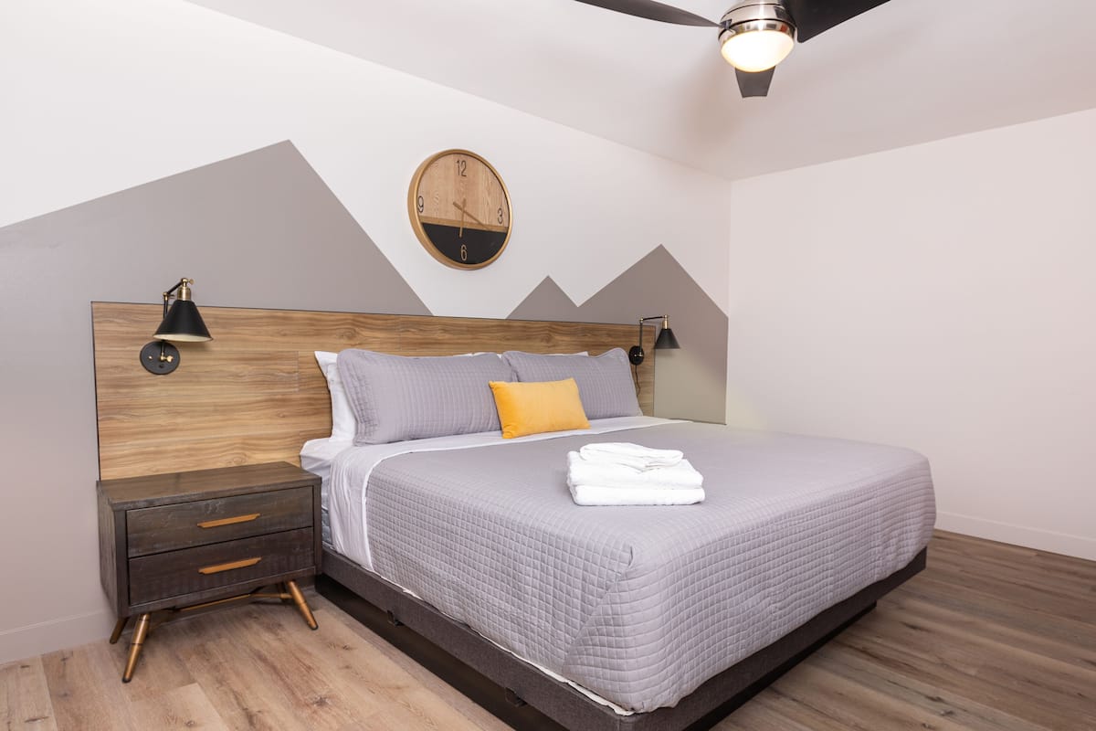 Une chambre à coucher moderne avec une tête de lit en bois, une horloge murale ronde, un ventilateur de plafond et un lit recouvert d’une couette et de coussins gris matelassés.